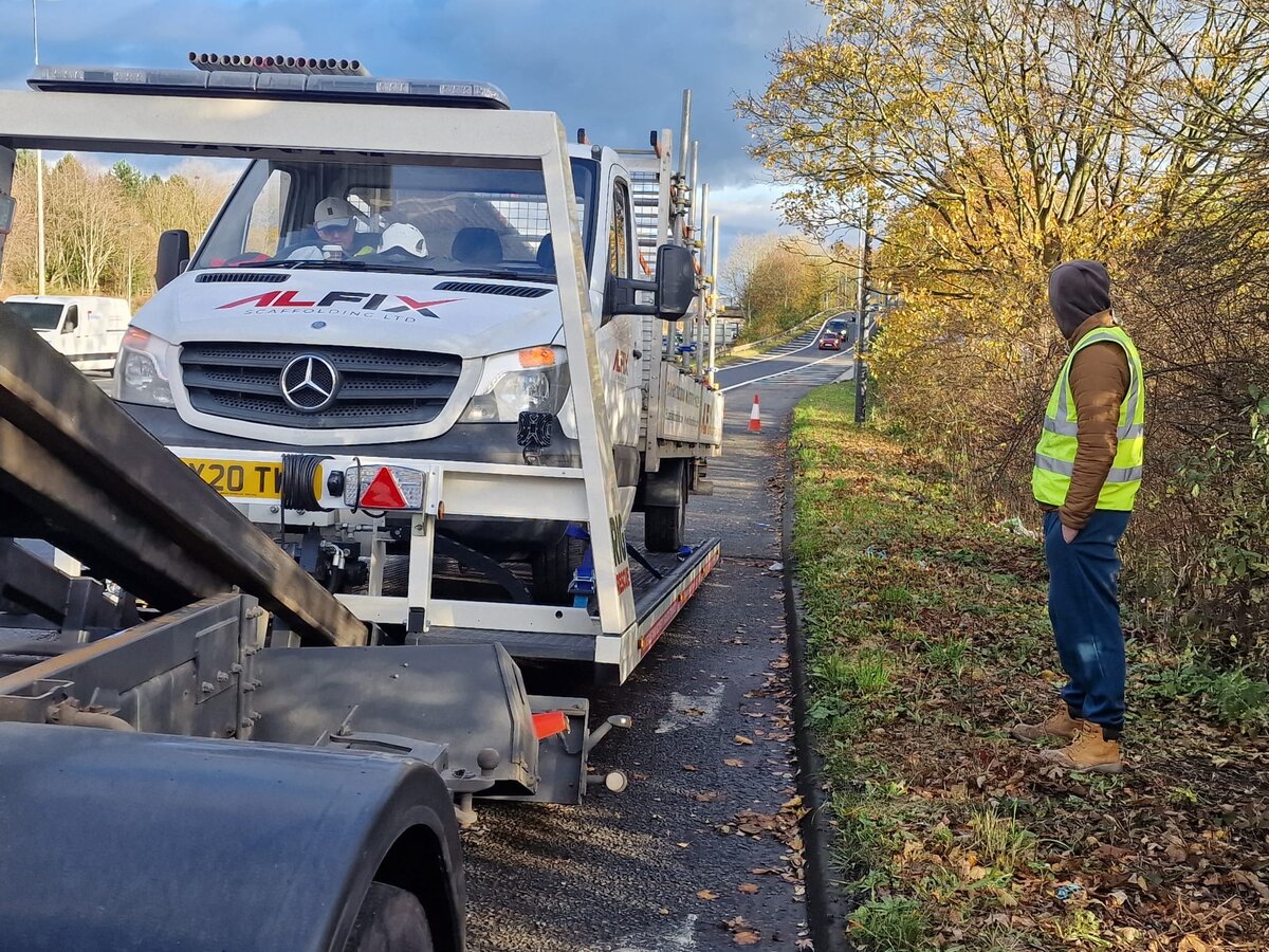 Alfix-branded Mercedes flatbed recovery truck loading a vehicle on a roadside in autumn with a Breakdown Man operator in high-vis stood beside it.