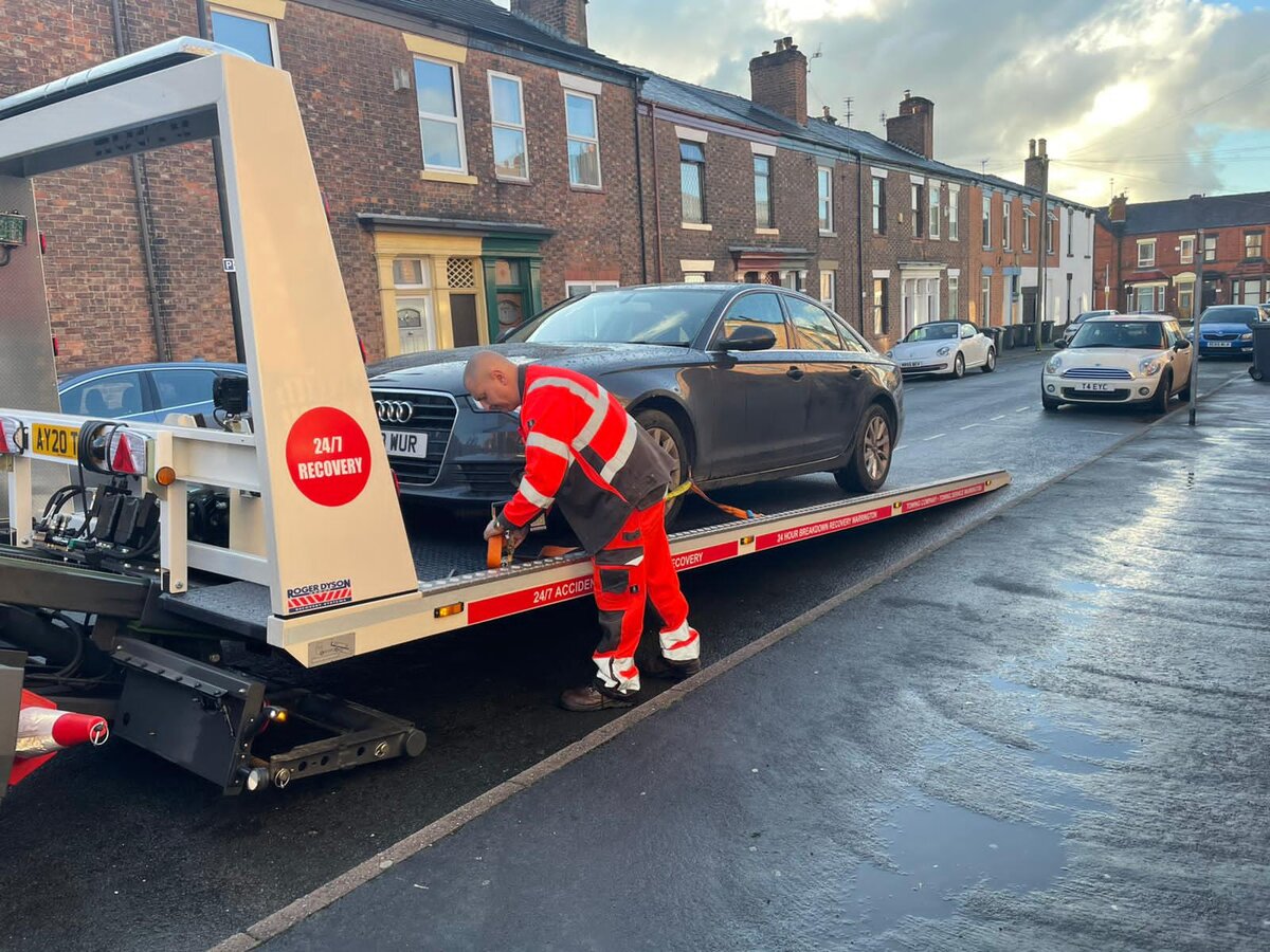 Breakdown Man operator in red high-vis crouched at the front wheel of a grey Audi A6 saloon as it loads onto the Breakdown Man flatbed on a Wigan terraced street