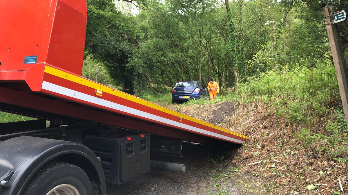 Red Breakdown Man flatbed recovery truck reversing onto a narrow rural lane to recover a blue saloon parked off the verge in the trees