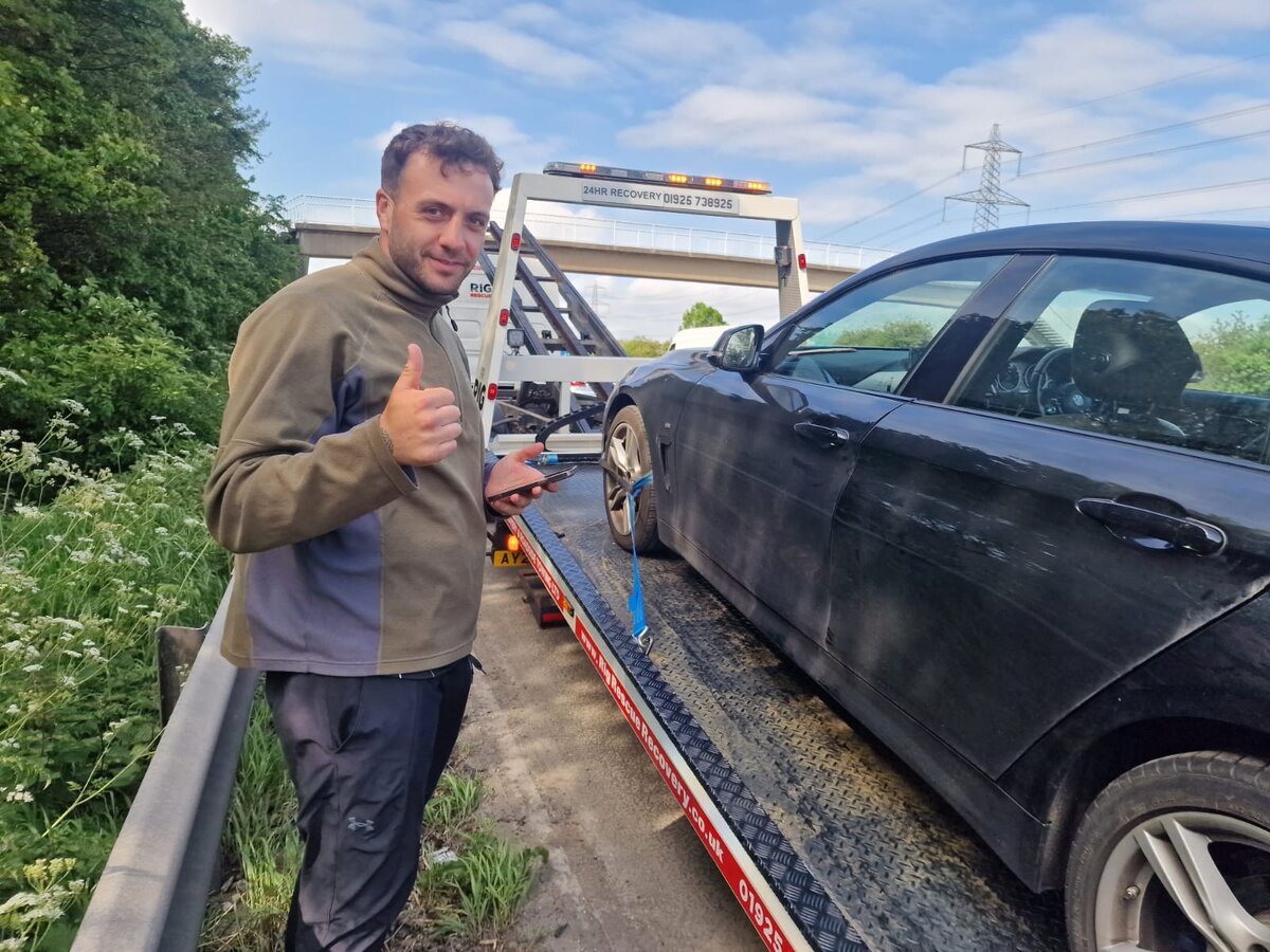 Black BMW saloon loaded on a Breakdown Man 24HR Recovery flatbed truck on a rural roadside verge with the recovery operator stood by giving a thumbs-up.