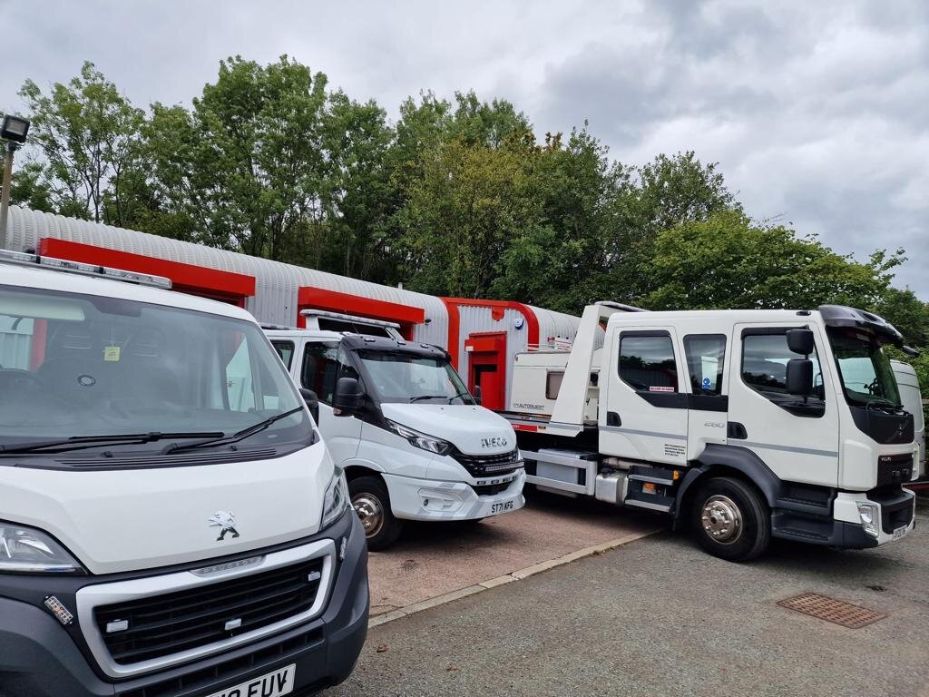 A Peugeot Boxer van, Iveco Daily van and DAF flatbed recovery truck parked side by side outside the Breakdown Man depot under cloudy skies.