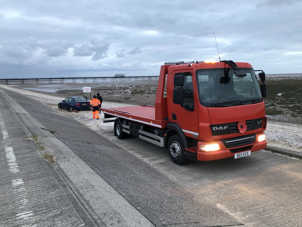 Red DAF LF flatbed recovery truck BE61 V25 parked along a coastal road with a pier in the distance, operator in high-vis attending a stranded car ahead