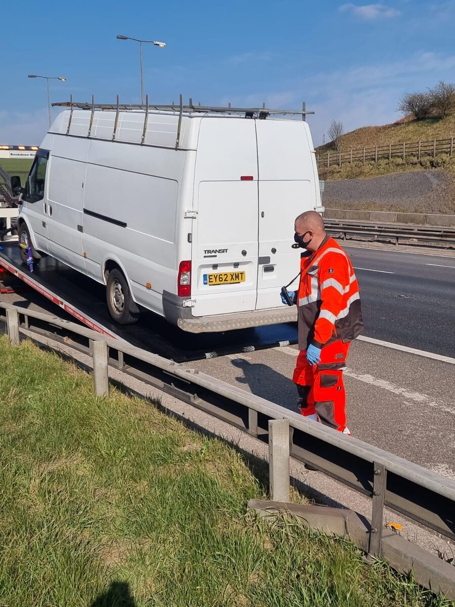 White Ford Transit van loaded onto Breakdown Man's flatbed beside the motorway, recovery operator in orange hi-vis checking the strap.