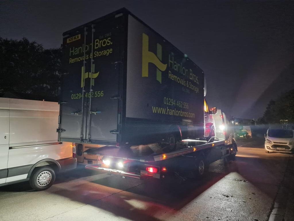 Hanlon Bros Removals & Storage Luton box truck loaded onto a Breakdown Man flatbed recovery truck at night, with a white van and other vehicles alongside.