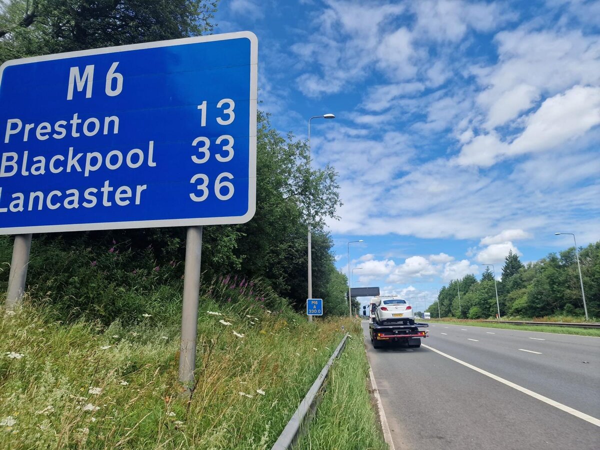 M6 motorway directional sign for Preston, Blackpool and Lancaster with a Breakdown Man flatbed recovery truck towing a vehicle along the carriageway in the background.