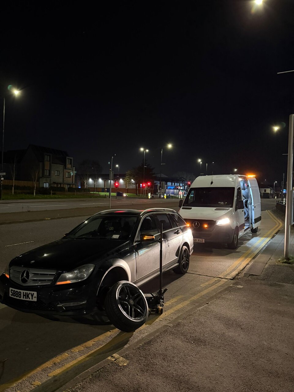 Black Mercedes saloon up on a trolley jack at night with the Breakdown Man recovery van parked behind, ready for a roadside tyre swap