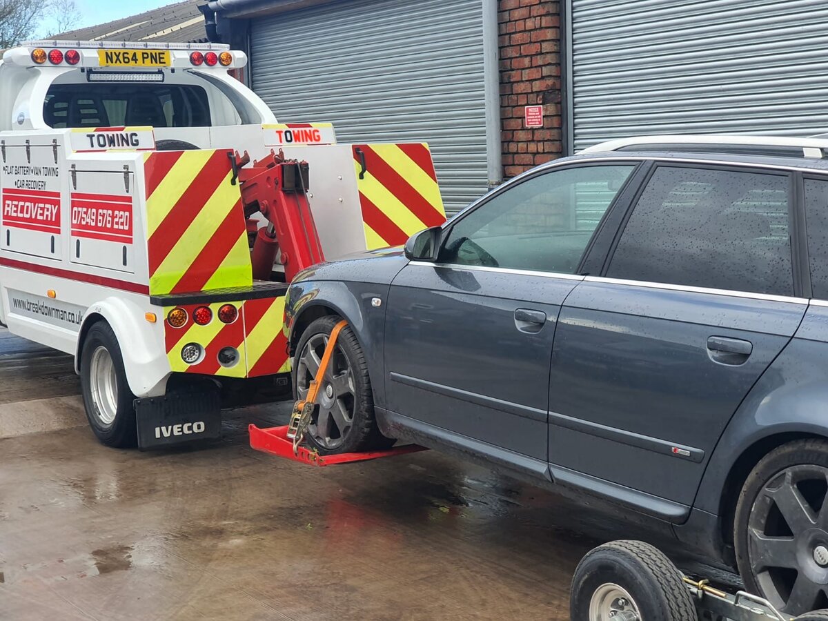 Breakdown Man recovery truck NX64 PNE towing a dark Audi A4 Avant outside an industrial unit with shutter doors