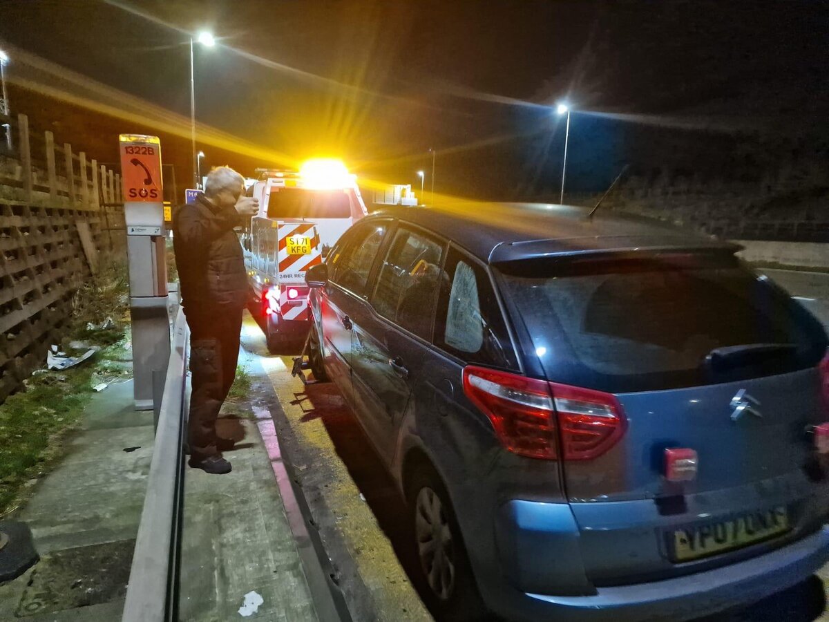 Silver Citroen people carrier broken down on the motorway hard shoulder near a numbered SOS phone, Breakdown Man recovery truck attending at night near Rochdale.