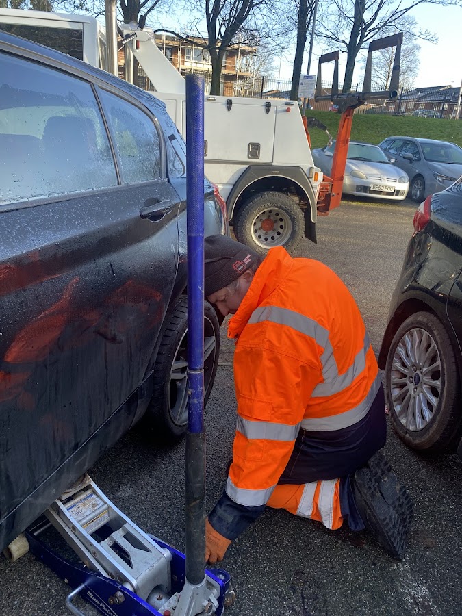 Simon from Breakdown Man in orange hi-vis kneeling beside a black hatchback in a car park, working a trolley jack to change a wheel