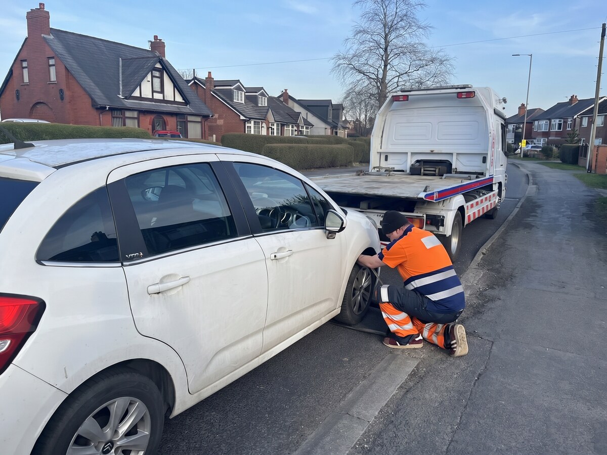Simon from Breakdown Man crouched at the front wheel of a white Citroen, fitting a replacement tyre on a residential street with the recovery truck parked behind