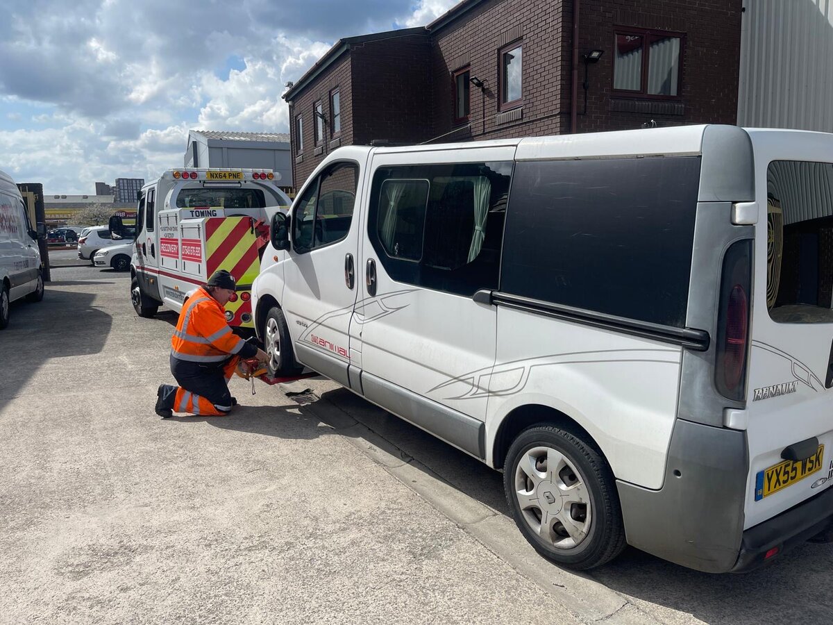 Simon from Breakdown Man kneeling beside a silver Renault Trafic van, fitting recovery wheel-strap restraints at the depot before transport