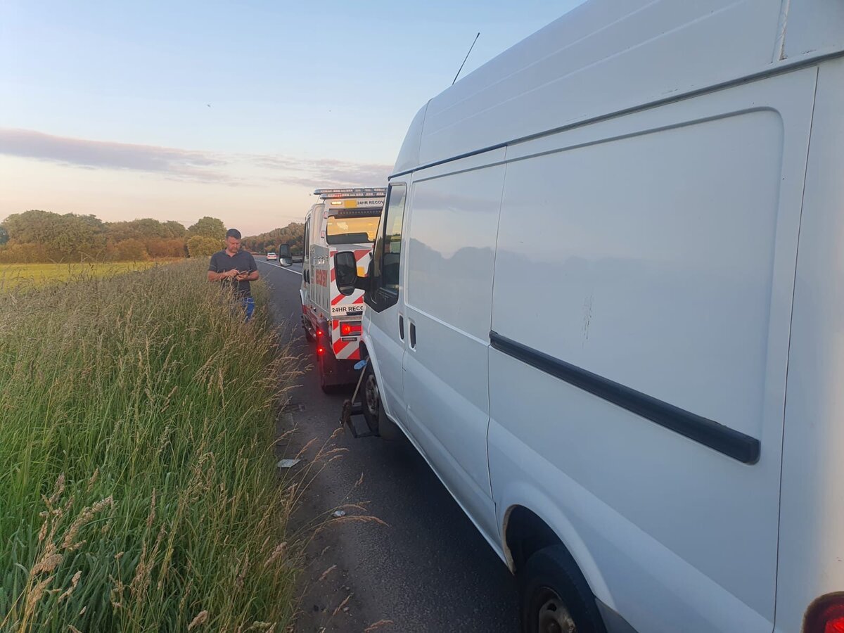 White Ford Transit van pulled over on a rural roadside verge at sunset with a Breakdown Man 24HR Recovery flatbed truck behind and the recovery operator stood by the van.