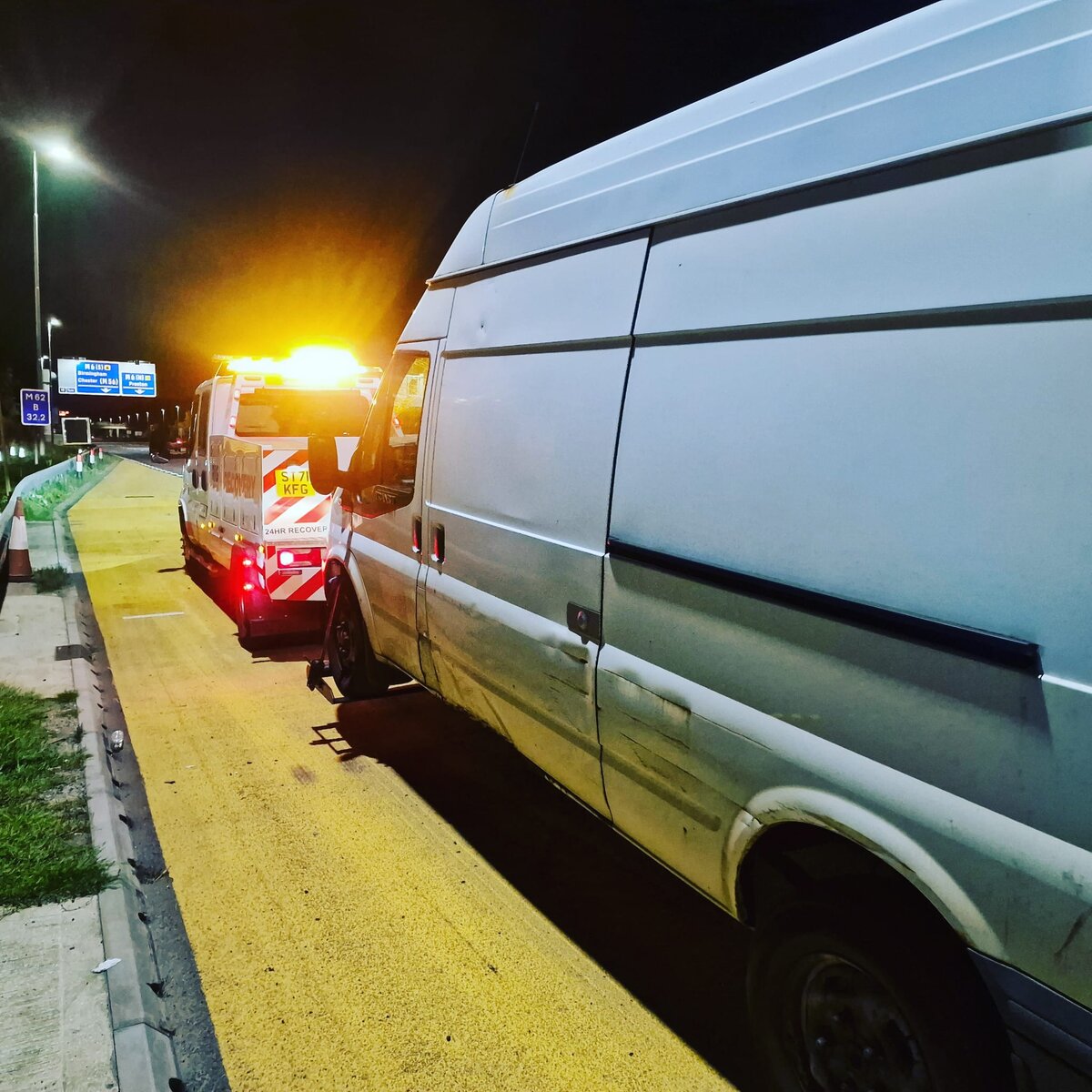 Silver Ford Transit van on the motorway slip road kerb at night with a Breakdown Man 24HR Recovery flatbed truck behind, blue motorway sign in the background.