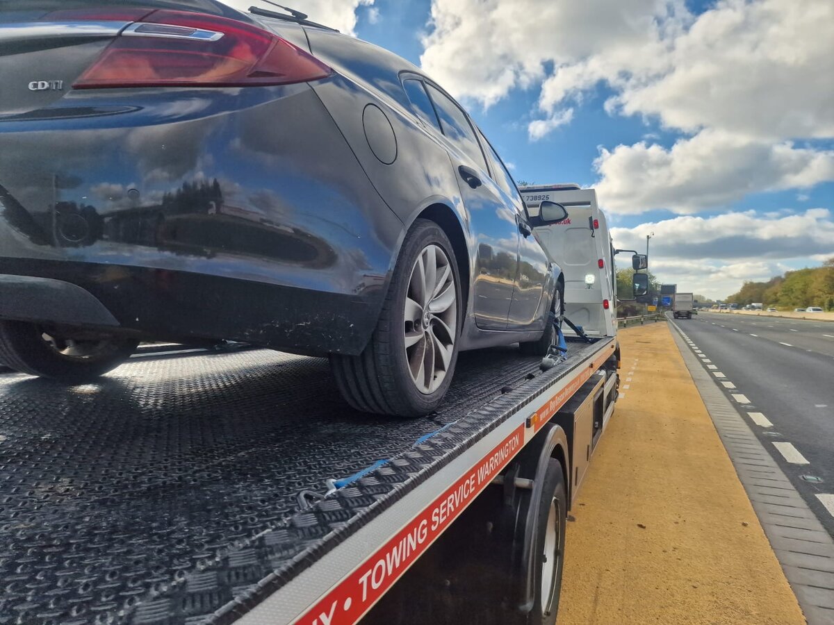 Black Vauxhall Insignia loaded on a Towing Service Warrington flatbed truck on the M62 hard shoulder under blue sky.